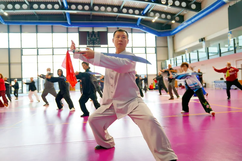 Tai chi sword form class practicing a low bow stance with straight swords in a training hall